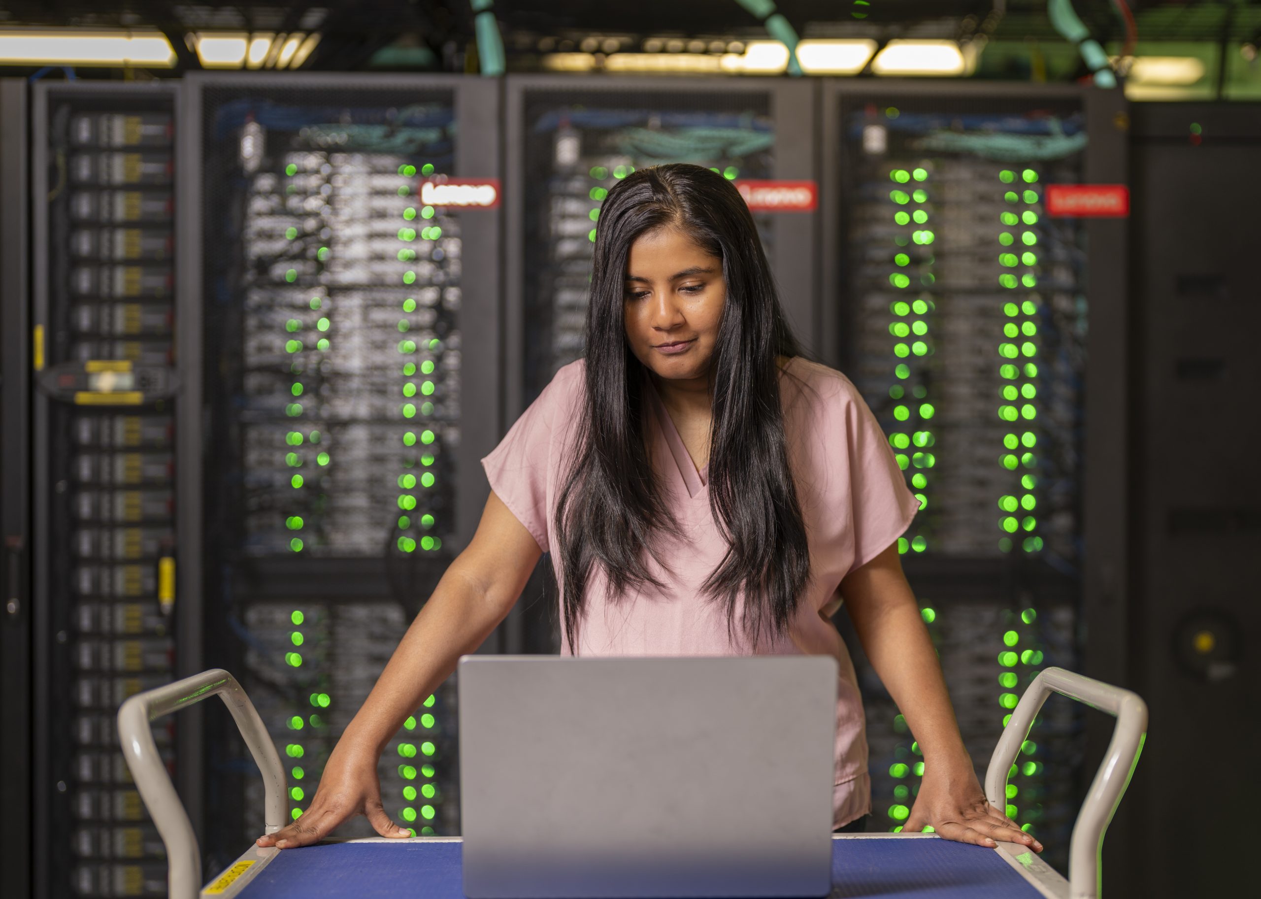Woman with long dark hair in a pink shirt, looking at a laptop screen, and standing in front of the National Computational Infrastructure super computer