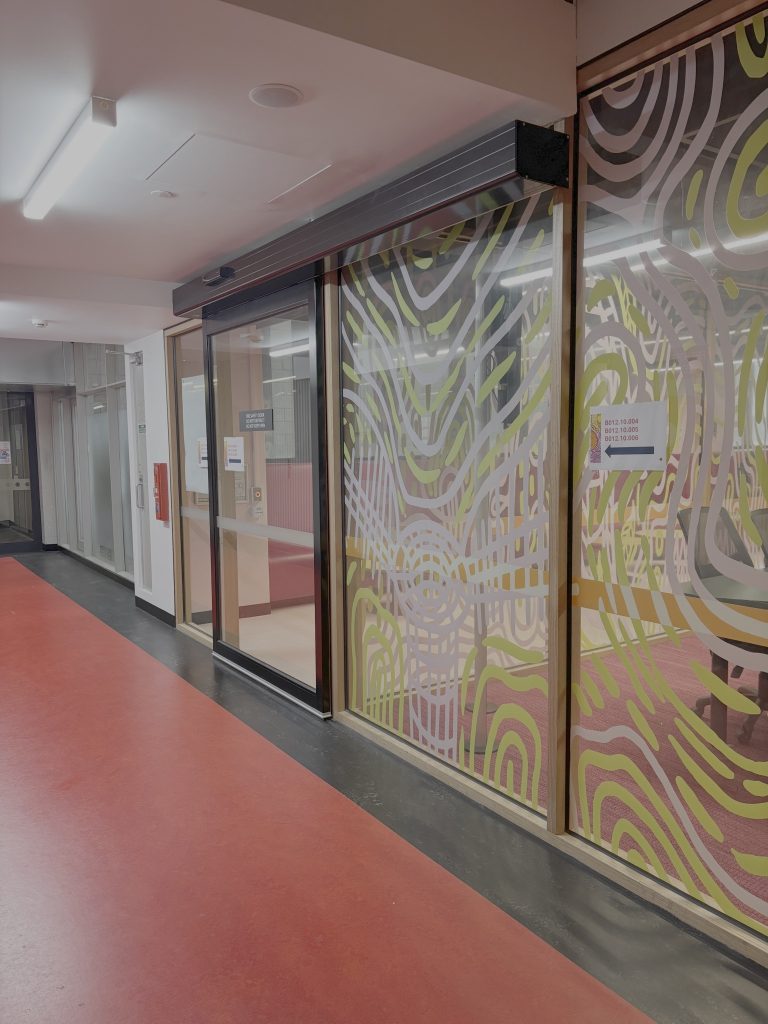 View of Room 4 entry, from the perspective of standing in the corridor outside the room. The room has a glass wall with yellow and white graphic art all over it, and a clear glass automatic sliding door to enter the space.
